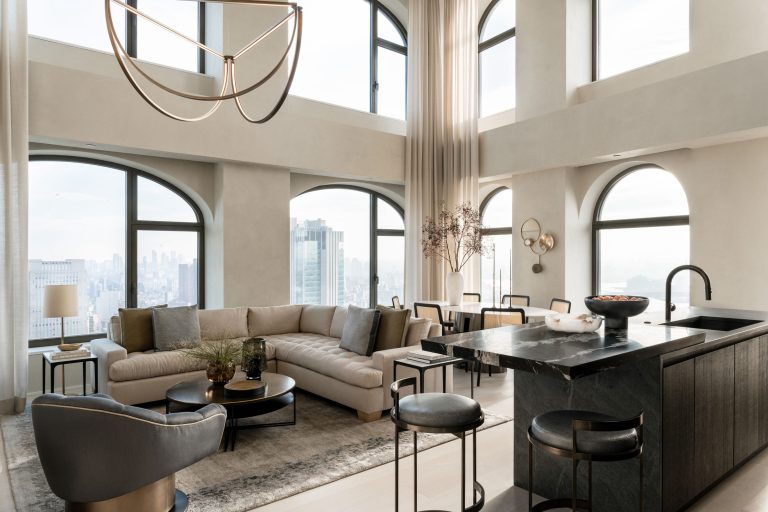A modern living room with high ceilings, large arched windows, and a city view. It features a beige sectional sofa, round coffee table, and gray rug. A sleek kitchen island with bar stools is on the right. A chandelier and tall curtains enhance the elegant, airy atmosphere.