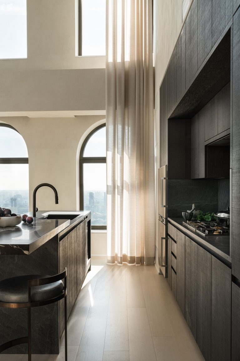 A modern kitchen with tall ceilings and large arched windows letting in natural light. It features dark wood cabinetry, a marble island with a black faucet, and high chairs. Light, sheer curtains partially cover the windows, creating a soft, elegant atmosphere.