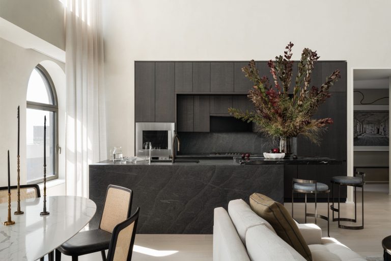 Modern kitchen with dark cabinets and a central island. A large floral arrangement sits atop the island. Natural light streams in through a tall arched window with sheer curtains. A dining table with chairs and a part of a sofa are visible, creating a sophisticated and cozy atmosphere.