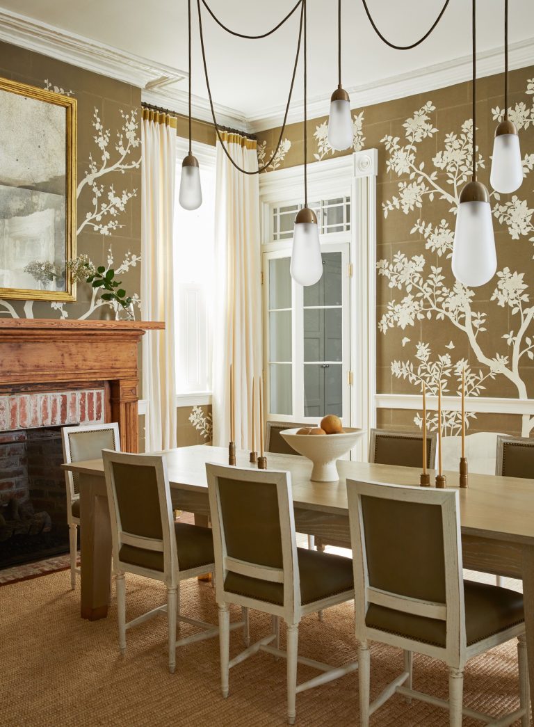 Elegant dining room with a light wood table and eight cushioned chairs. The walls feature a brown floral pattern. A wooden fireplace mantel with a painting above it, large windows with cream curtains, and modern pendant lights hang from the ceiling. A decorative bowl centers the table.