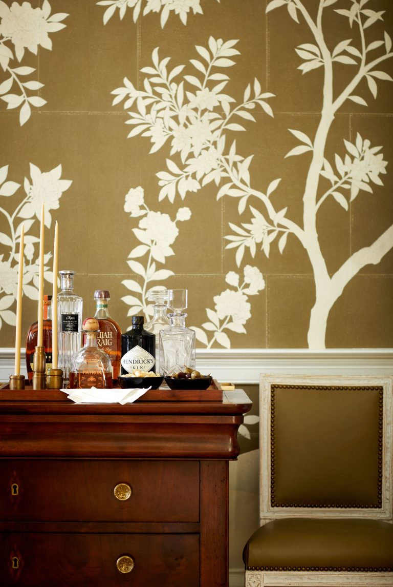 A vintage wooden cabinet with various liquor bottles, including a bottle of gin and whiskey, and glasses sits against a wall with a decorative white floral pattern on olive background. A tray holds a note and candlesticks. A green upholstered chair is partially visible.