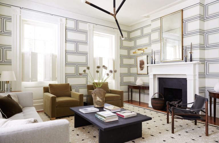 A modern living room with geometric wallpaper. It features a light gray sofa, two brown armchairs, a black coffee table with books, and a black fireplace. A large mirror hangs above the mantel, and unique lighting fixtures are present. A patterned rug covers the floor.