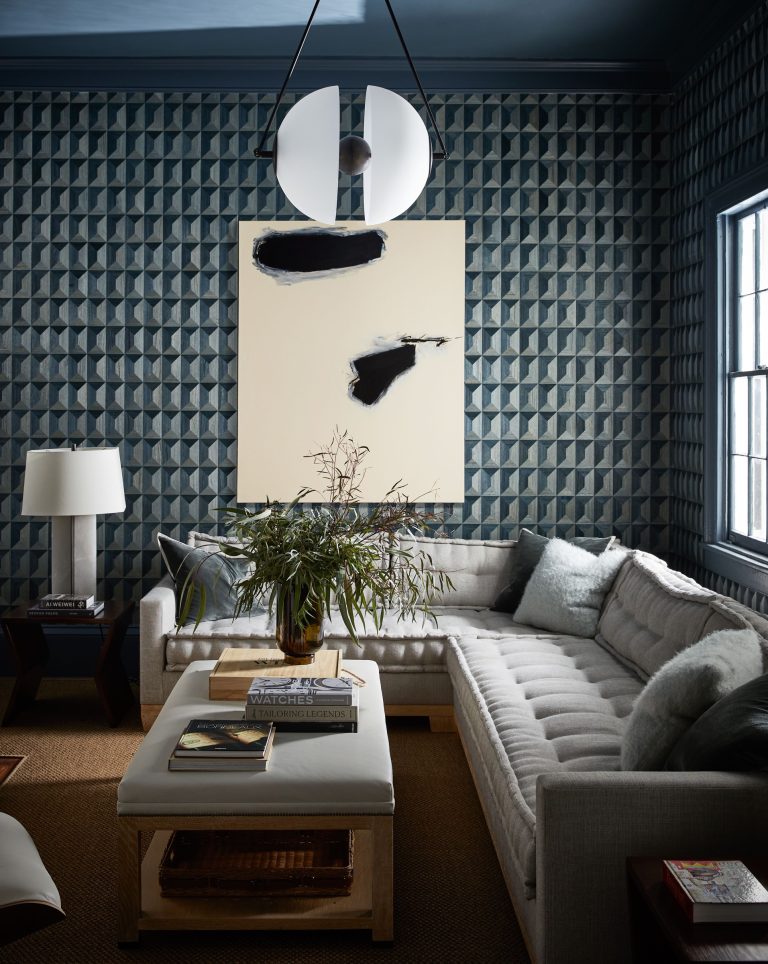 A modern living room with geometric-patterned blue walls and a gray sectional sofa. A large abstract painting with black brushstrokes hangs above the sofa. A white ottoman with books and a plant sits in the center. A circular, dual-color pendant light hangs from the ceiling.