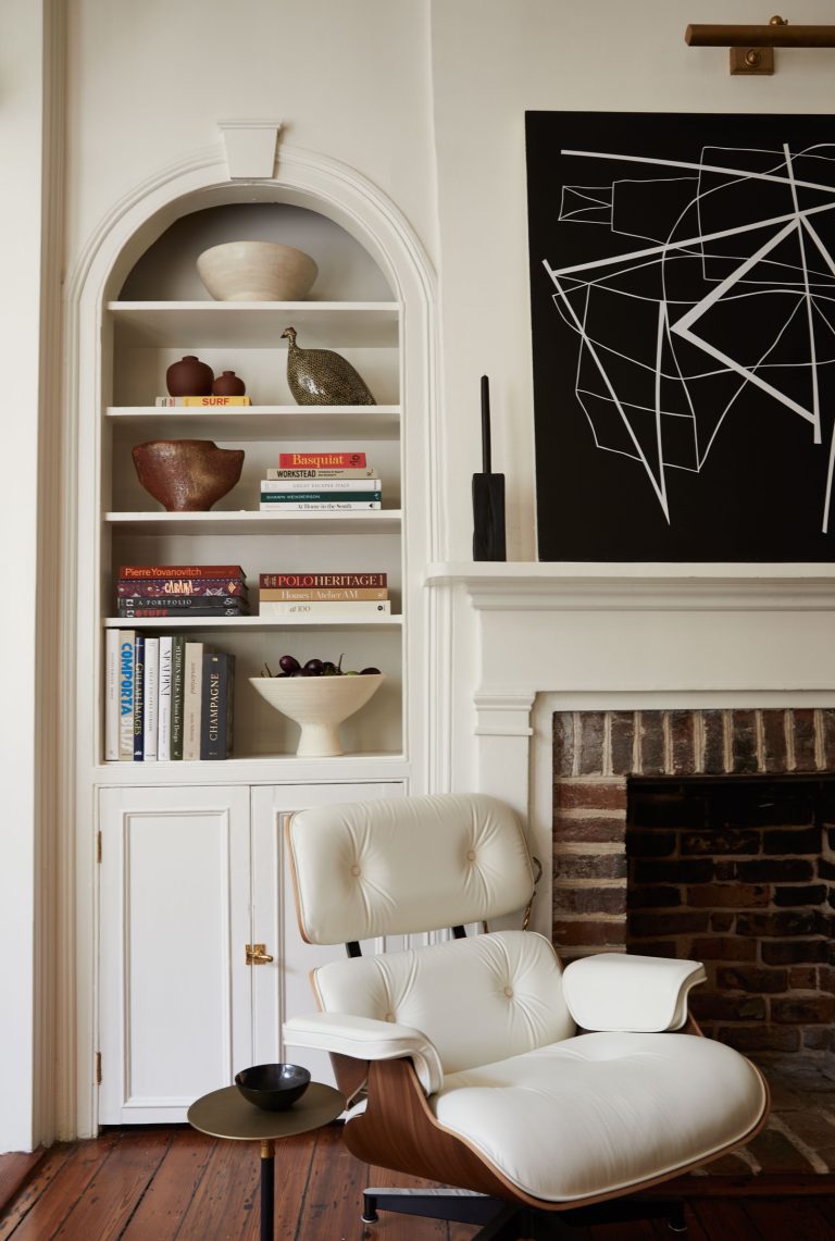 A cozy living room features a white Eames lounge chair and ottoman on a wooden floor. To the left, a white built-in bookshelf displays books, ceramic vases, and a peacock figurine. A fireplace with a brick interior is topped by a black geometric art piece.