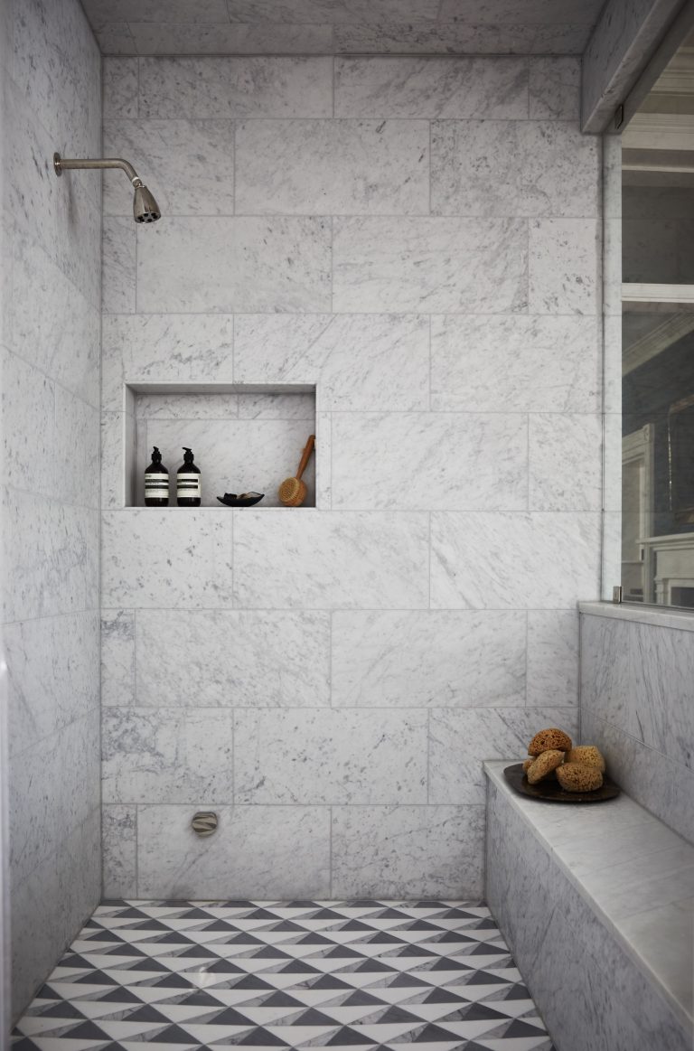 A minimalist shower with white marble tiles and a geometric patterned floor. Theres a niche in the wall holding bottles and a brush. A small silver showerhead is mounted on the left. The shower has a marble bench on the right with a couple of sponges placed on it.