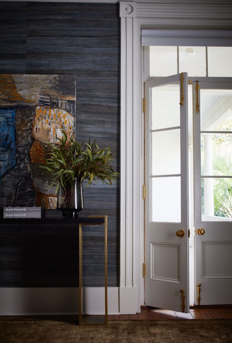 Stylish room with a dark textured wall featuring an abstract painting above a slim black and gold table. A vase with greenery sits on the table alongside stacked books. Double glass doors with brass handles lead to a sunlit patio, contrasting with the rooms darker tones.
