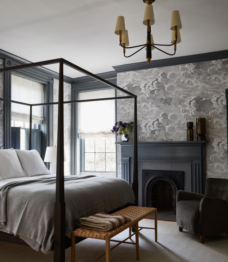 A cozy bedroom features a black four-poster bed with white and gray linens. A wicker bench sits at the foot of the bed. The room has gray patterned wallpaper, a fireplace with a mirror above, a gray armchair, and a modern chandelier. Light streams in through two large windows.