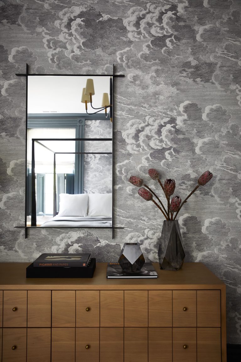 A modern interior features a wooden dresser topped with books and a geometric vase holding dried flowers. Above, a rectangular mirror reflects a four-poster bed and a wall lamp. The wallpaper displays a gray and white cloud pattern, adding texture to the space.