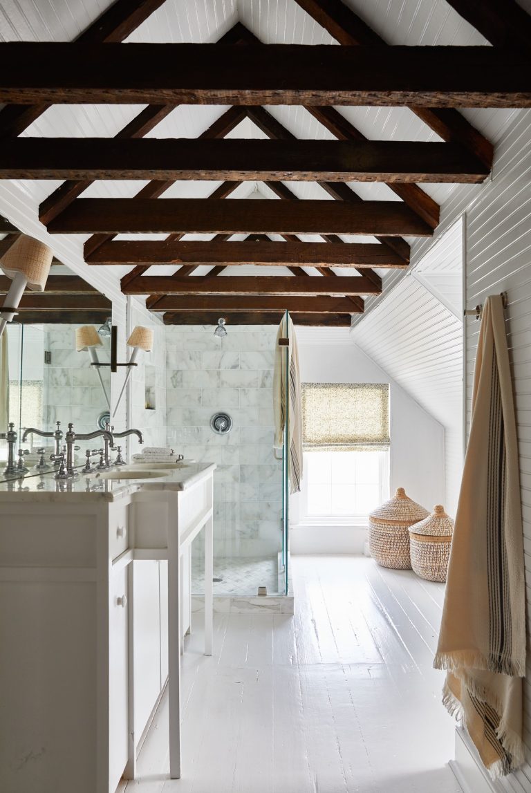 A rustic bathroom with exposed wooden beams on a white ceiling. White walls and floor complement the space. A glass shower with marble tiles stands on the left, next to a white vanity with dual sinks and wall-mounted lamps. Two woven baskets and a window with a woven shade are on the right.