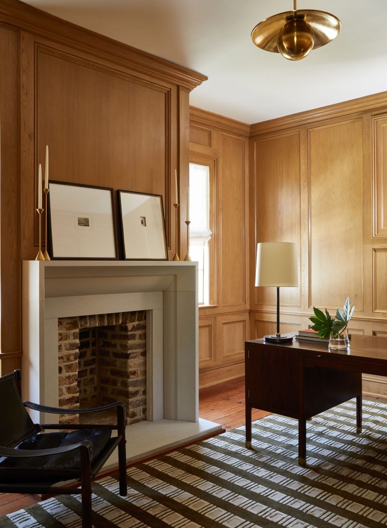 A cozy room features a wooden paneled interior with a modern fireplace. Two framed artworks on the mantel, a dark wooden desk with a table lamp and plant, and a black chair complement the space. The striped rug and brass ceiling light add warmth and elegance to the decor.
