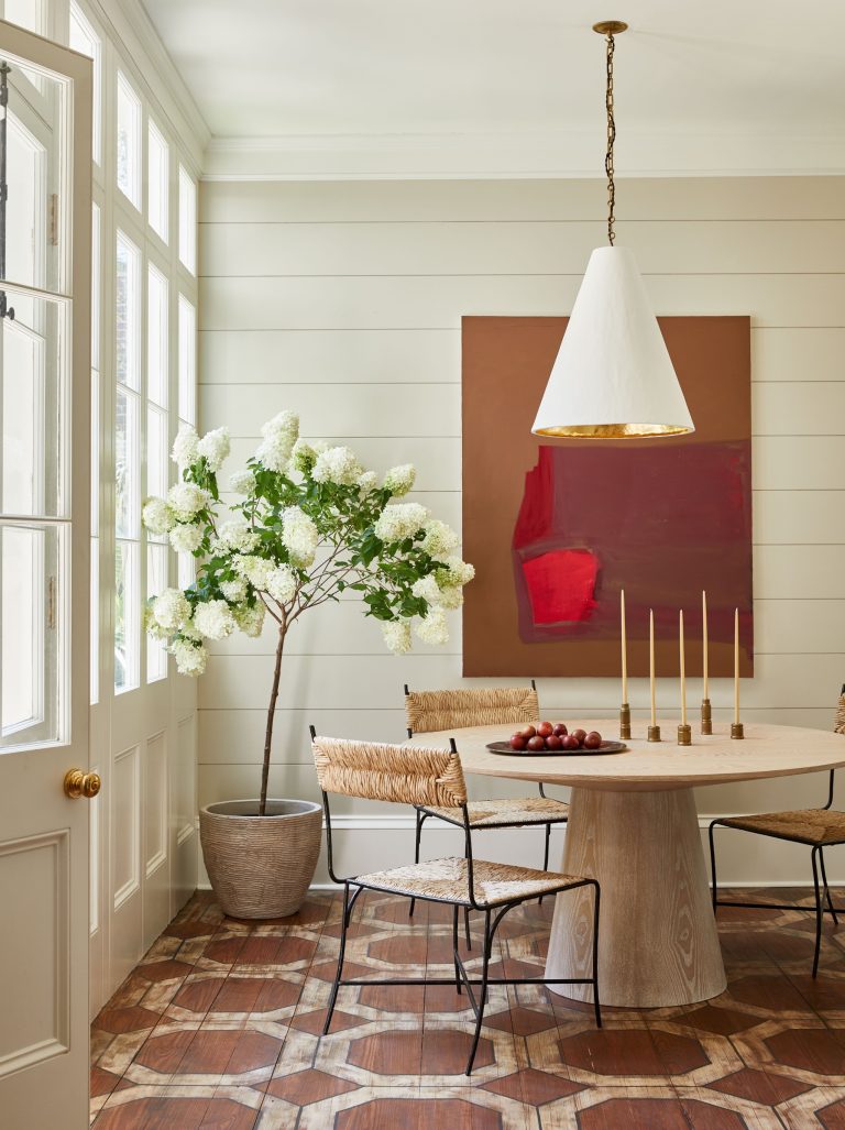 A dining area with a light wood round table and four wicker chairs on a patterned wood floor. A large potted plant with white blooms is beside a window. A white cone-shaped pendant light hangs above the table, which is set with a bowl of red apples and tall candlesticks. A large abstract painting with red and brown tones hangs on the wall.