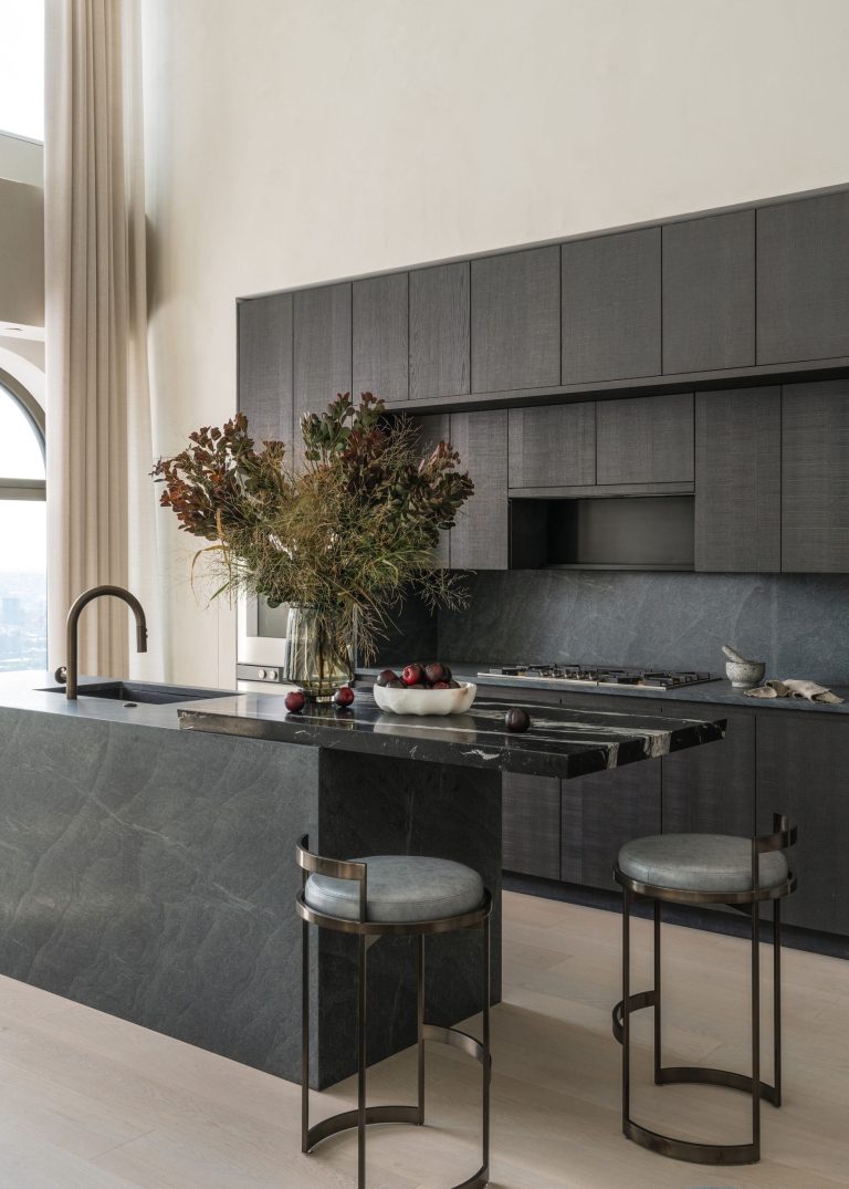 Modern kitchen with dark gray cabinetry and a marble island. Two round stools with metal frames sit at the island. A large vase holds dried floral arrangements atop the counter, alongside a bowl of apples. Tall windows with light curtains allow natural light to fill the space.