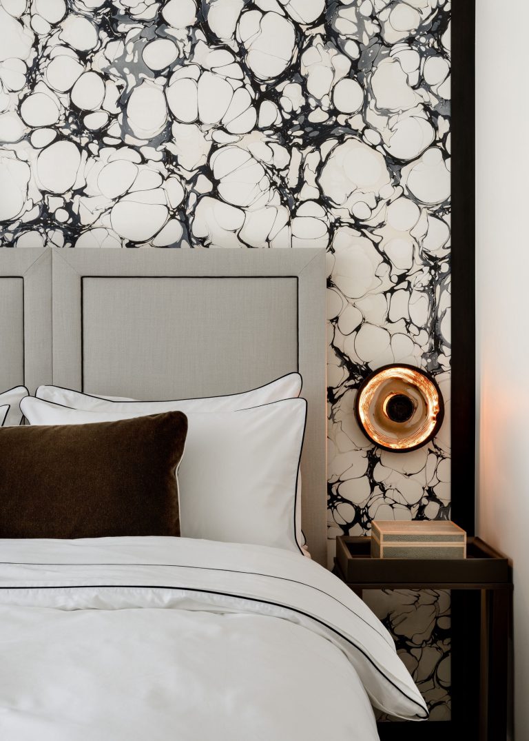 A modern bedroom featuring a light gray upholstered headboard against a black and white marbled wall. The bed is dressed with white linens, bordered pillows, and a brown accent pillow. A brass wall sconce emits warm light, and a small bedside table holds a stacked book.
