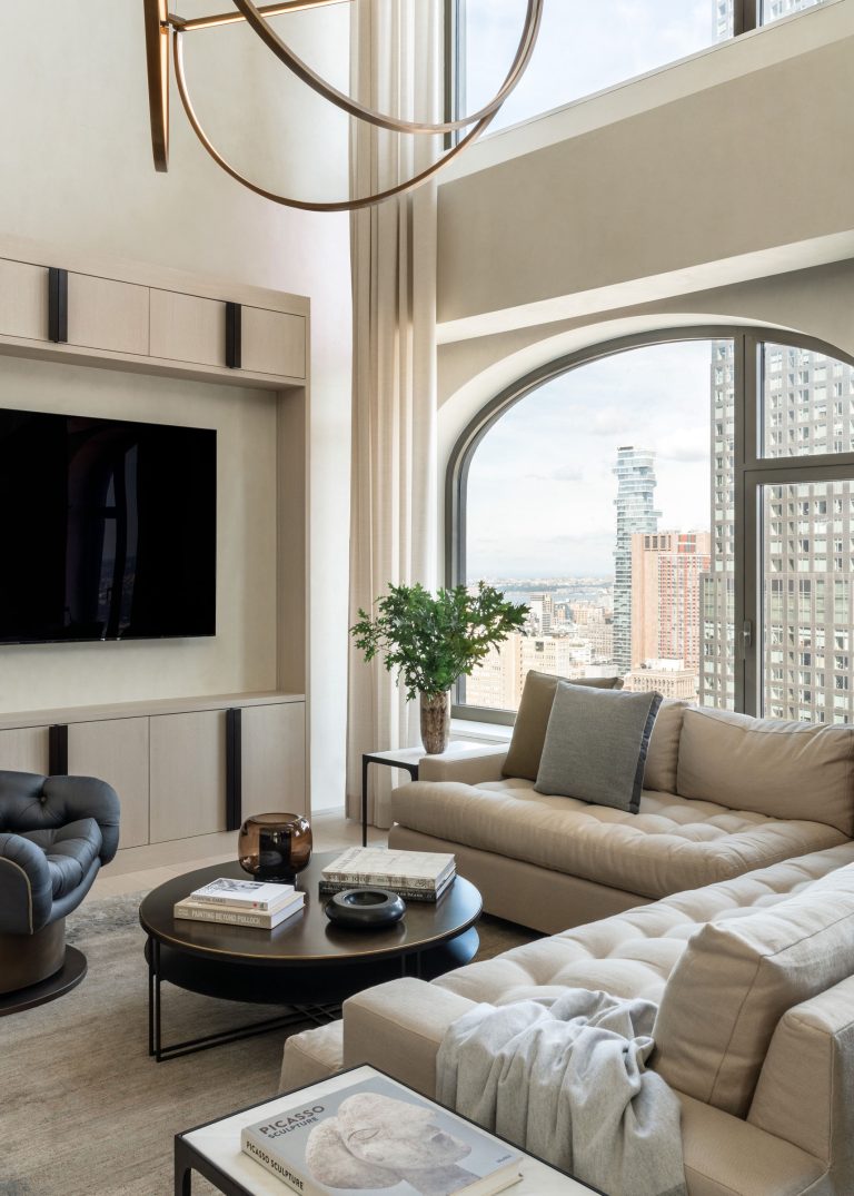 A modern living room with large windows overlooking a city skyline. It features a beige sectional sofa, a black round coffee table with books and a brown vase, a gray armchair, and a built-in media unit with a large flat-screen TV. A potted plant adds a touch of greenery.