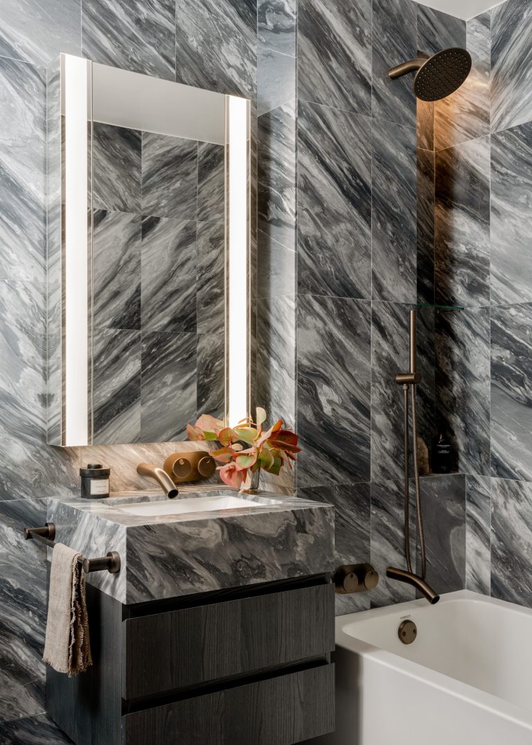A modern bathroom with marble-patterned gray tiles on the walls. A rectangular mirror with built-in lighting hangs above a dark stone vanity. A faucet and sink sit below, adorned with a small vase of foliage. A towel is draped over a nearby rack. The shower features a round, black showerhead.