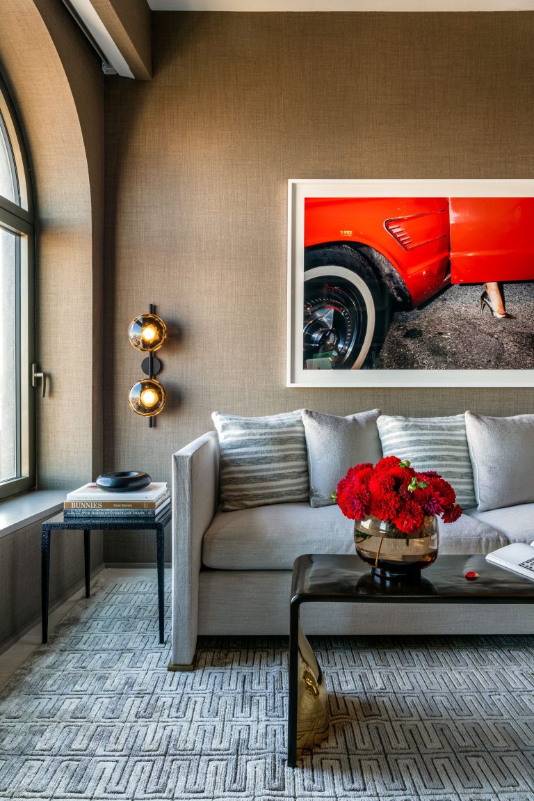 A modern living room with a gray sofa adorned with striped cushions. A black coffee table holds a bouquet of red flowers. The wall features a large photo of a vintage red car and a womans foot. Two spherical wall lights flank the artwork. A geometric-patterned rug covers the floor.