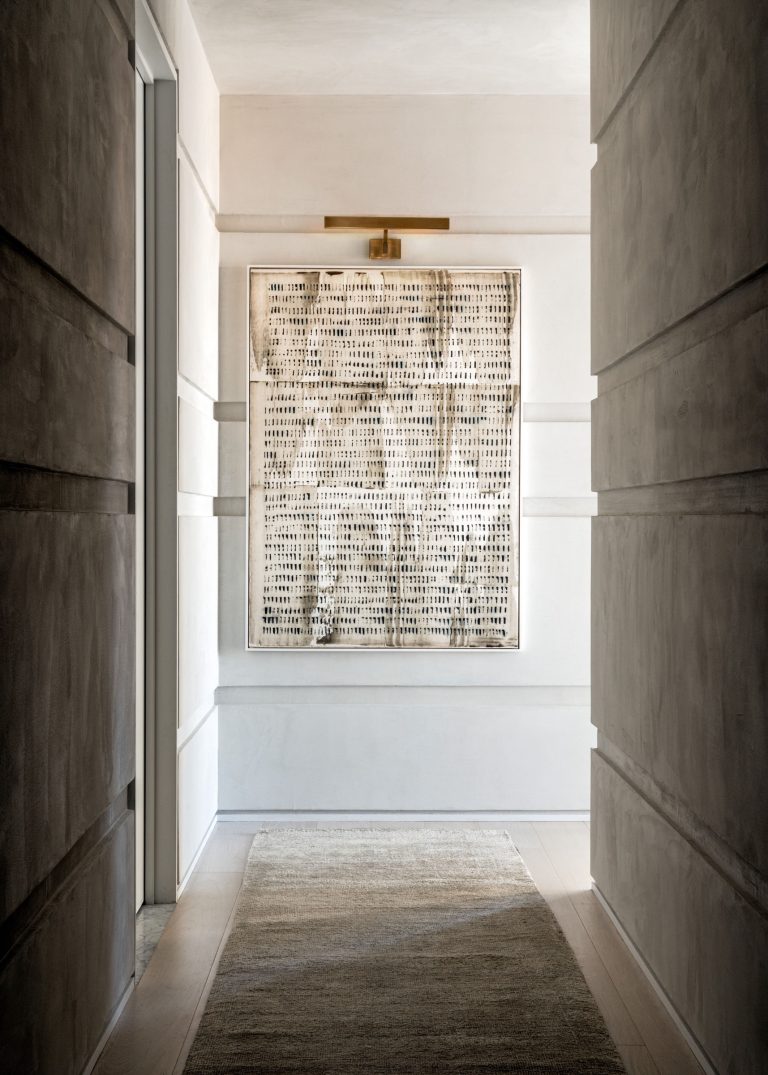 A minimalist hallway with textured concrete walls leading to a piece of abstract art framed and mounted on the far wall. The artwork features a grid of small black marks on a white background. A wall-mounted light above illuminates the art. A neutral-toned rug covers the wooden floor.