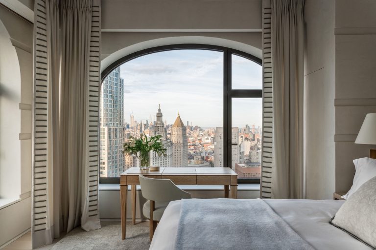 A modern bedroom features a large arched window with a cityscape view, including skyscrapers. The room has a neatly made bed with white linens, a wooden desk, a chair, and a potted plant. Curtains frame the window, and soft natural light fills the space.