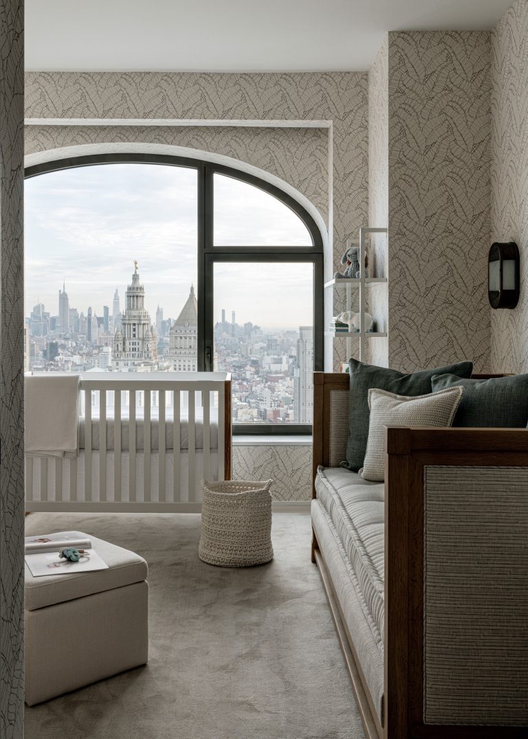Stylish nursery with a crib overlooking a city skyline through a large arched window. The room features neutral tones, a patterned wallpaper, a cushioned bench with green and white pillows, a woven basket, and a small, rectangular ottoman, creating a serene and modern atmosphere.