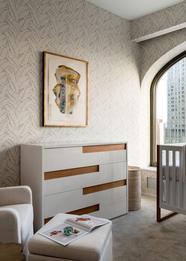A modern nursery with a white crib and dresser featuring wooden accents. The room has a large arched window and abstract wallpaper. A framed abstract art piece hangs on the wall. A cozy armchair with a footstool holds baby items. A woven basket is placed near the dresser.