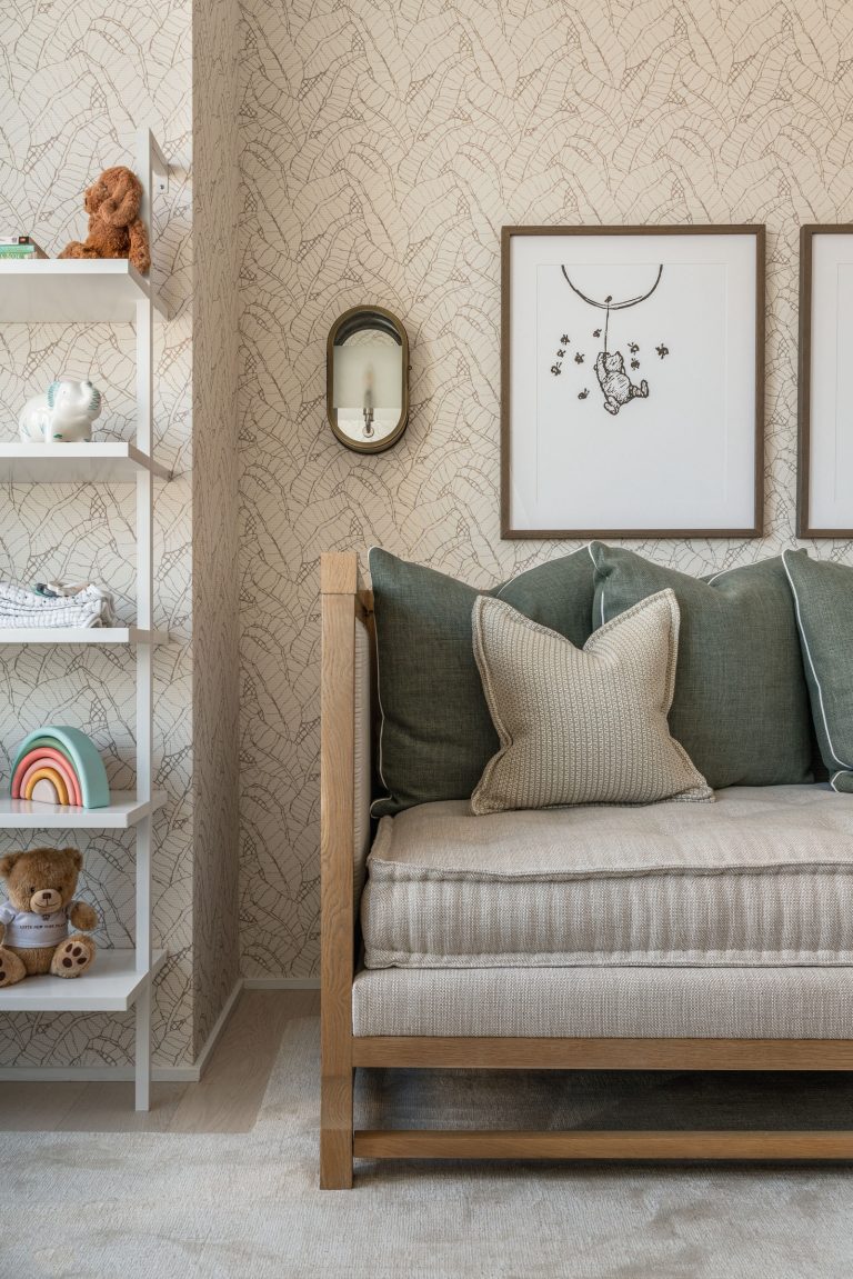 A cozy living room corner features a beige sofa with green and beige pillows. Two framed illustrations hang on a textured wallpaper. A white shelf on the left holds decorative items, including a teddy bear, a ceramic elephant, and a rainbow. A wall-mounted sconce adds soft light.