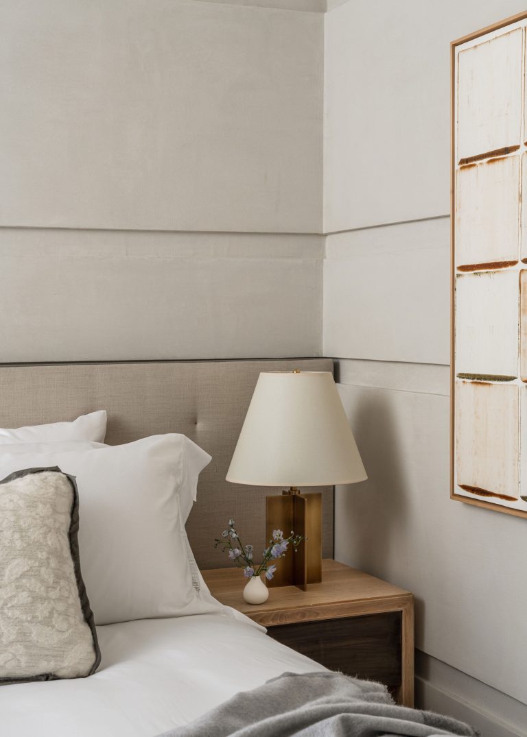 A neatly arranged bedroom corner features a light gray padded headboard and white bedding with patterned pillows. A wooden nightstand holds a modern lamp with a white shade and a small vase with blue flowers. A piece of abstract artwork is partially visible on the wall.