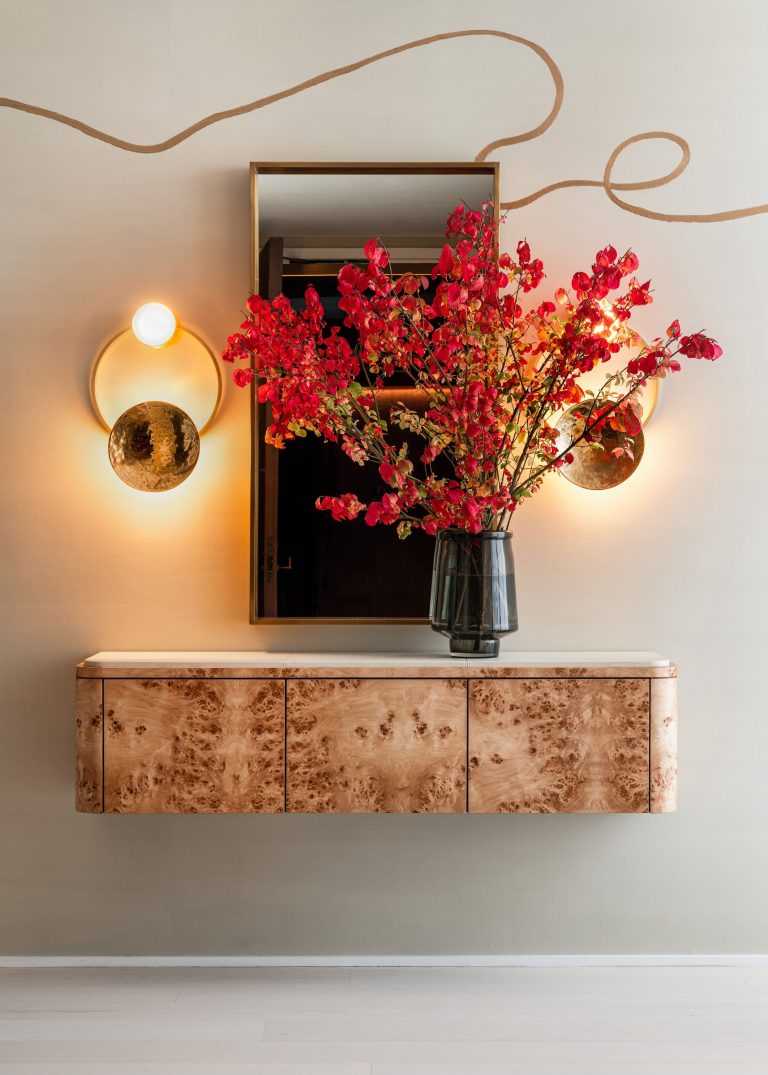 A modern interior features a wall-mounted wooden cabinet with a white top. Above it hangs a mirror flanked by two round, glowing wall lights. A black vase holds vibrant red flowers, and a brown abstract line decorates the light beige wall. The floor is light wood.