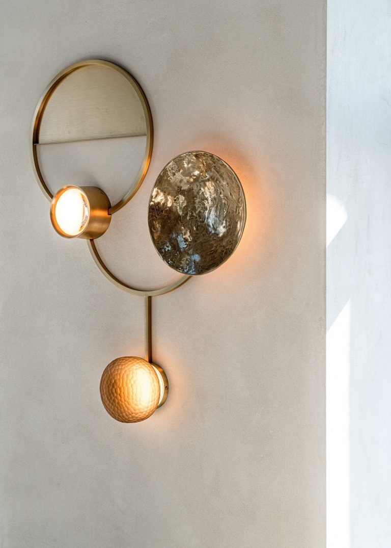 Modern wall sconce with a circular design on a beige wall. It has two lights: one enclosed in a metal dome with a textured surface, and the other in a small, downward-facing, smooth, gold-toned sphere. A partial shadow is cast on the wall, and sunlight is visible from a nearby window.