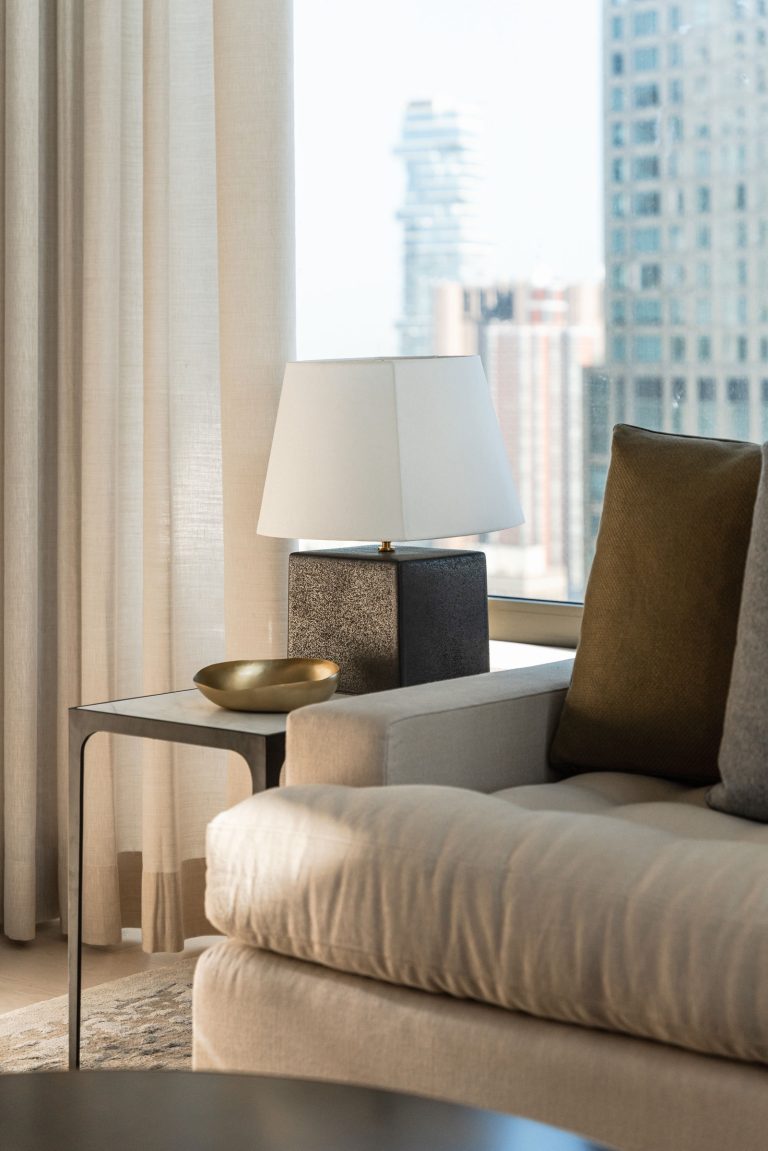 A modern living room with a beige sofa featuring dark and light brown cushions. A sleek metal side table holds a textured lamp with a white shade and a small gold bowl. Sheer curtains frame a large window with a blurry cityscape view in the background.