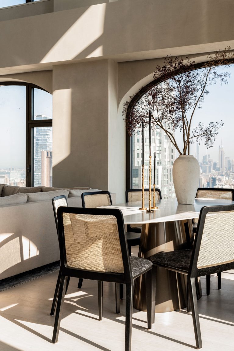 A modern dining room with large arched windows showcasing a city skyline. A round table with dark chairs surrounds a vase with branches. Soft sunlight illuminates the room, highlighting a neutral-toned sofa in the background. The cityscape adds a striking backdrop.